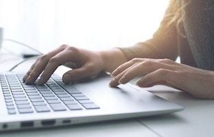Person typing on a laptop with a bright window in the background. The lighting creates a softly lit scene, and the focus is on the hands actively working on the keyboard.